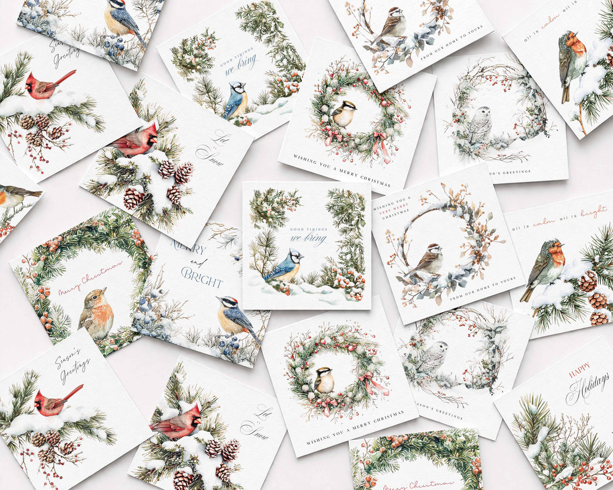 A flat-lay arrangement of multiple white Christmas greeting cards featuring watercolor illustrations of birds, wreaths, pine branches, and holiday text, photographed on a pale pink background.