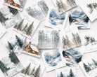 A flat-lay arrangement of multiple watercolor winter landscape Christmas cards scattered on a light background. The prints depict snowy forests, mountains, cabins, and rivers in soft blue, gray, white, and brown tones, with some cards featuring handwritten text and small holiday greetings.
