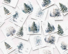 A flat-lay arrangement of multiple Christmas cards featuring watercolor winter trees and snowy forest scenes in soft blue, gray, and green tones on white backgrounds.