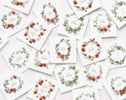 A flat-lay arrangement of multiple white square Christmas cards scattered on a light background, each printed with a watercolor floral wreath and holiday greeting text.