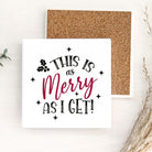 A square white decorative sign with the phrase “THIS IS as Merry AS I GET!” in black and dark pink lettering, shown in front of a cork board on a light wood surface with dried grass and small green leaves as props.