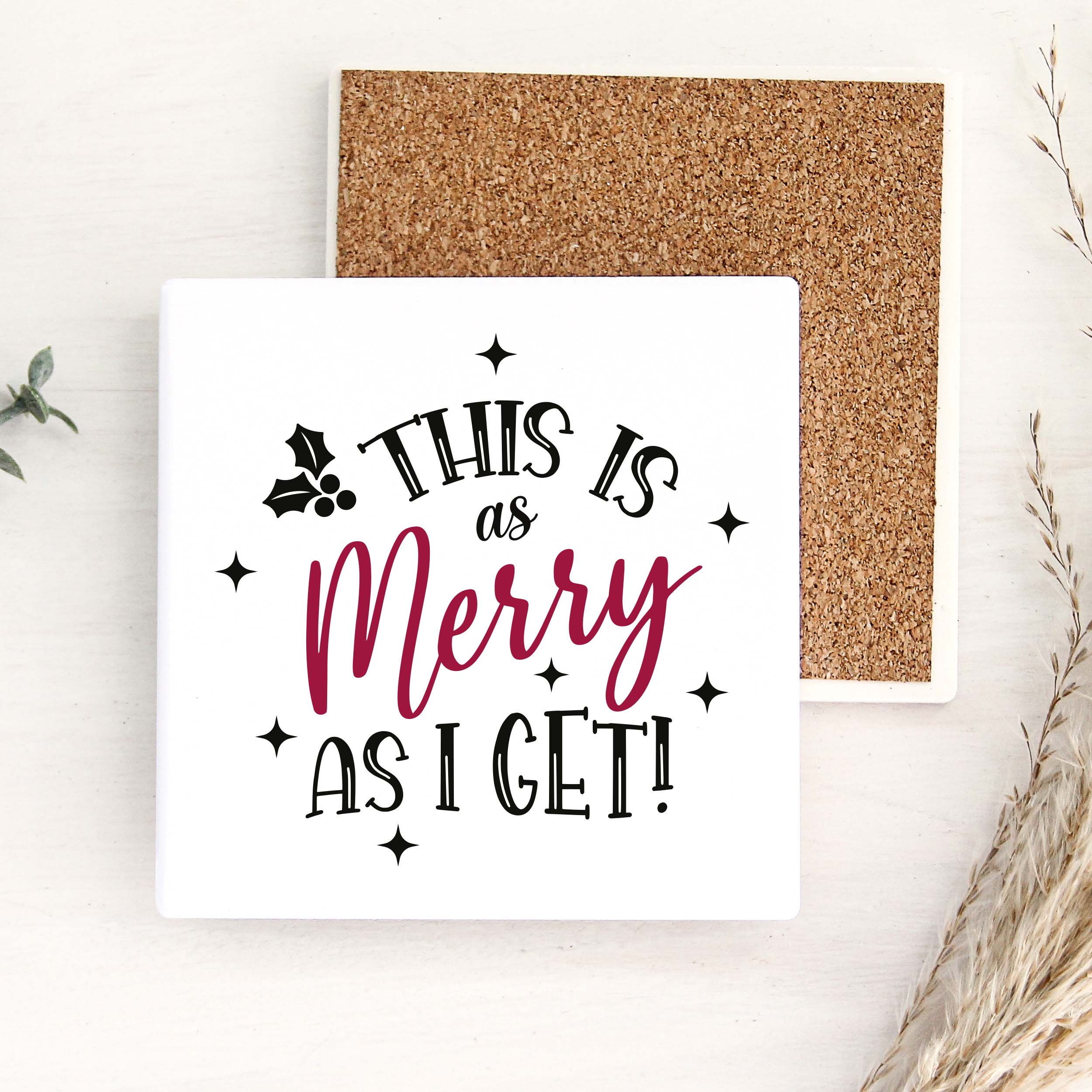 A square white decorative sign with the phrase “THIS IS as Merry AS I GET!” in black and dark pink lettering, shown in front of a cork board on a light wood surface with dried grass and small green leaves as props.