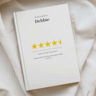 A white hardcover notebook or journal lying on draped cream fabric, printed with a review template that says 'You left a review for: Debbie' above a row of yellow stars and a short review comment.