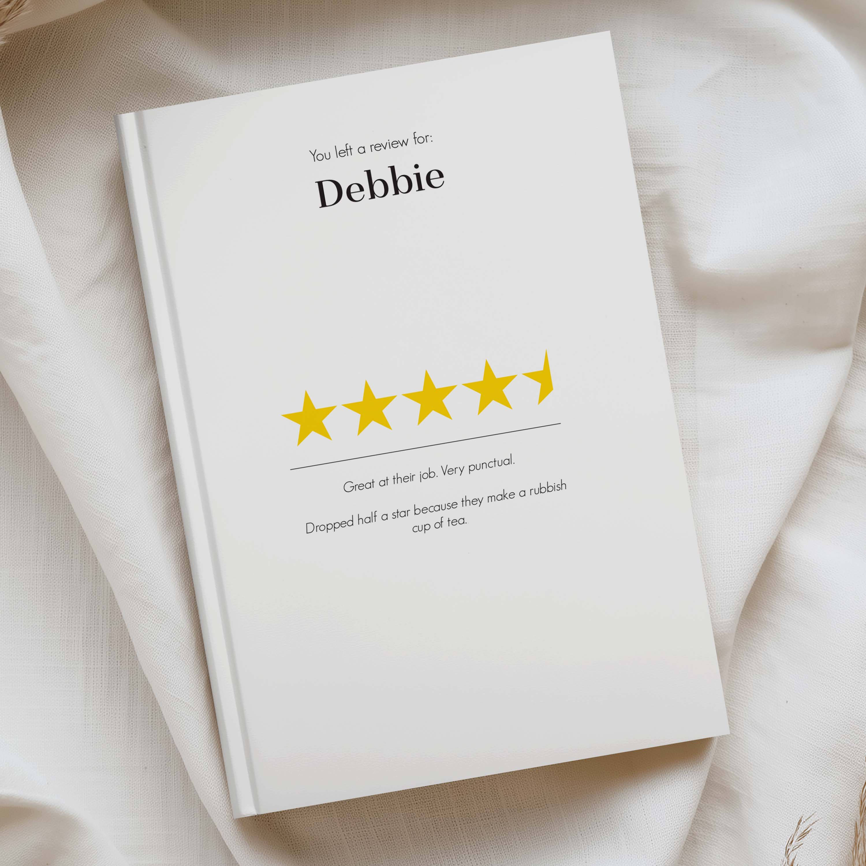 A white hardcover notebook or journal lying on draped cream fabric, printed with a review template that says 'You left a review for: Debbie' above a row of yellow stars and a short review comment.