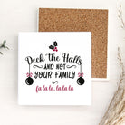 A square white decorative sign with a Christmas-themed quote reading 'Deck the Halls and not your family fa la la, la la la' in black and pink lettering, shown in front of a cork board on a light wood surface with dried grass and small green leaves as props.