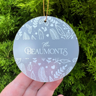 A round frosted glass Christmas ornament with white botanical patterns and the text 'The Beaumonts' in the center, held in a hand against a background of green pine branches.