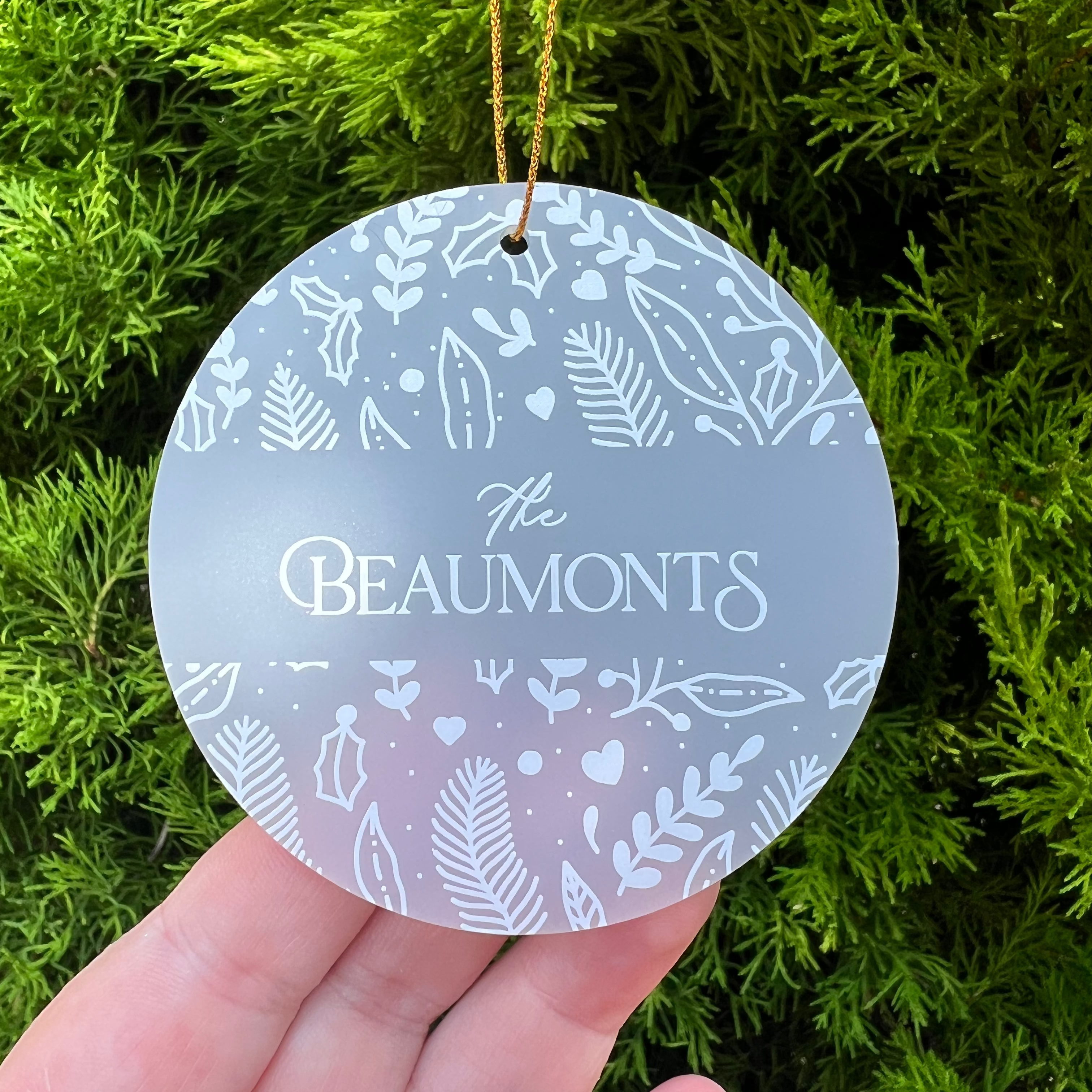 A round frosted glass Christmas ornament with white botanical patterns and the text 'The Beaumonts' in the center, held in a hand against a background of green pine branches.