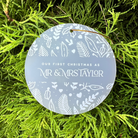 A round frosted glass Christmas ornament with white botanical illustrations and the text 'OUR FIRST CHRISTMAS AS MR & MRS TAYLOR', displayed on a bed of bright green pine needles.
