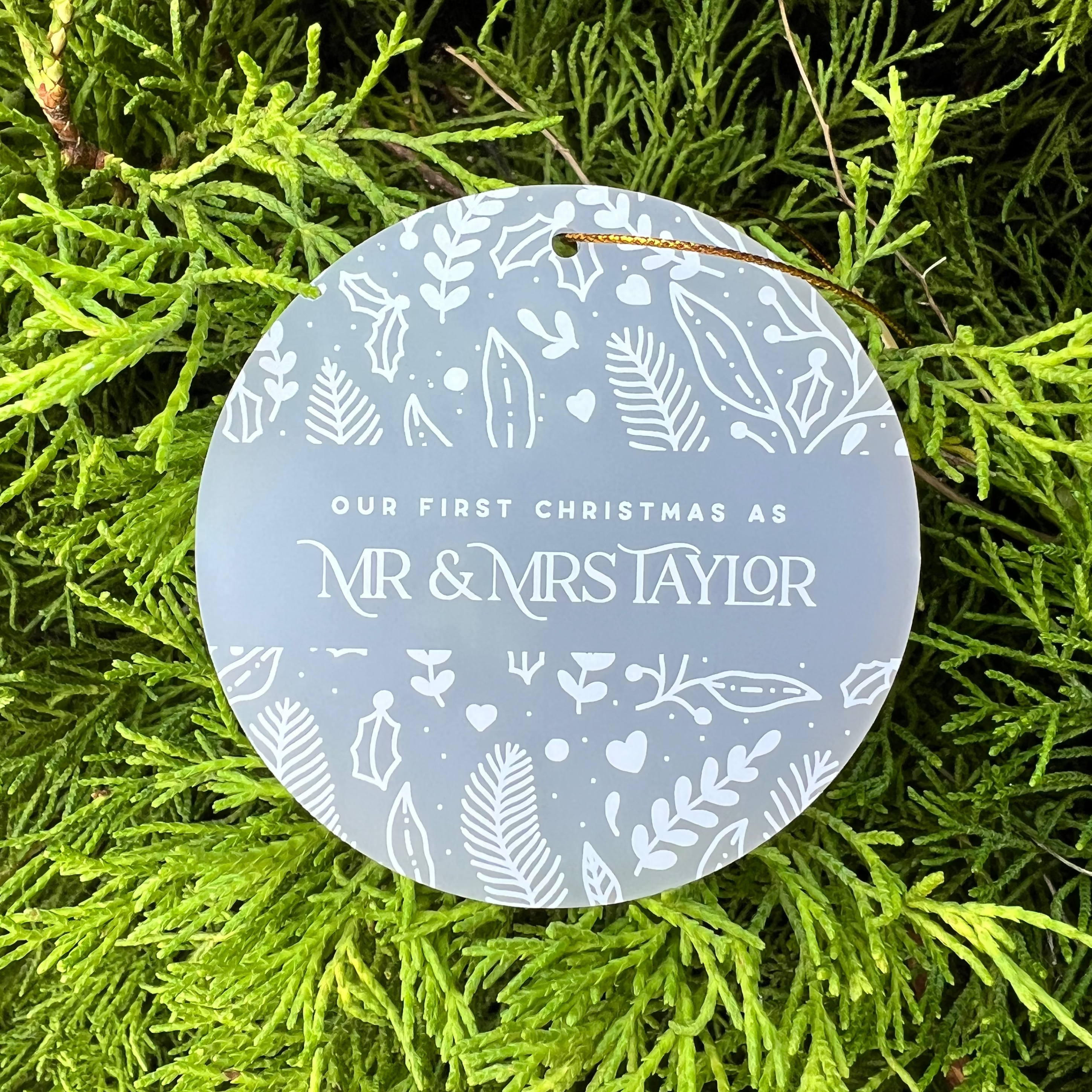 A round frosted glass Christmas ornament with white botanical illustrations and the text 'OUR FIRST CHRISTMAS AS MR & MRS TAYLOR', displayed on a bed of bright green pine needles.