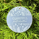 A round personalized Christmas ornament with a frosted light blue-gray finish and white botanical illustrations, hanging from a brown string in green foliage. The ornament is personalized with the text 'CHARLOTTE'S first christmas'.