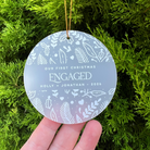 A round personalized Christmas ornament held in a hand against a background of green pine branches. The ornament is frosted light gray with white leaf and heart patterns and reads 'OUR FIRST CHRISTMAS ENGAGED HOLLY + JONATHAN - 2026'.