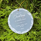 A round frosted glass Christmas ornament with white floral border and personalized text reading 'Our First Christmas at 27 Harbour Street' and names, hanging from a gold string amidst bright green pine branches.