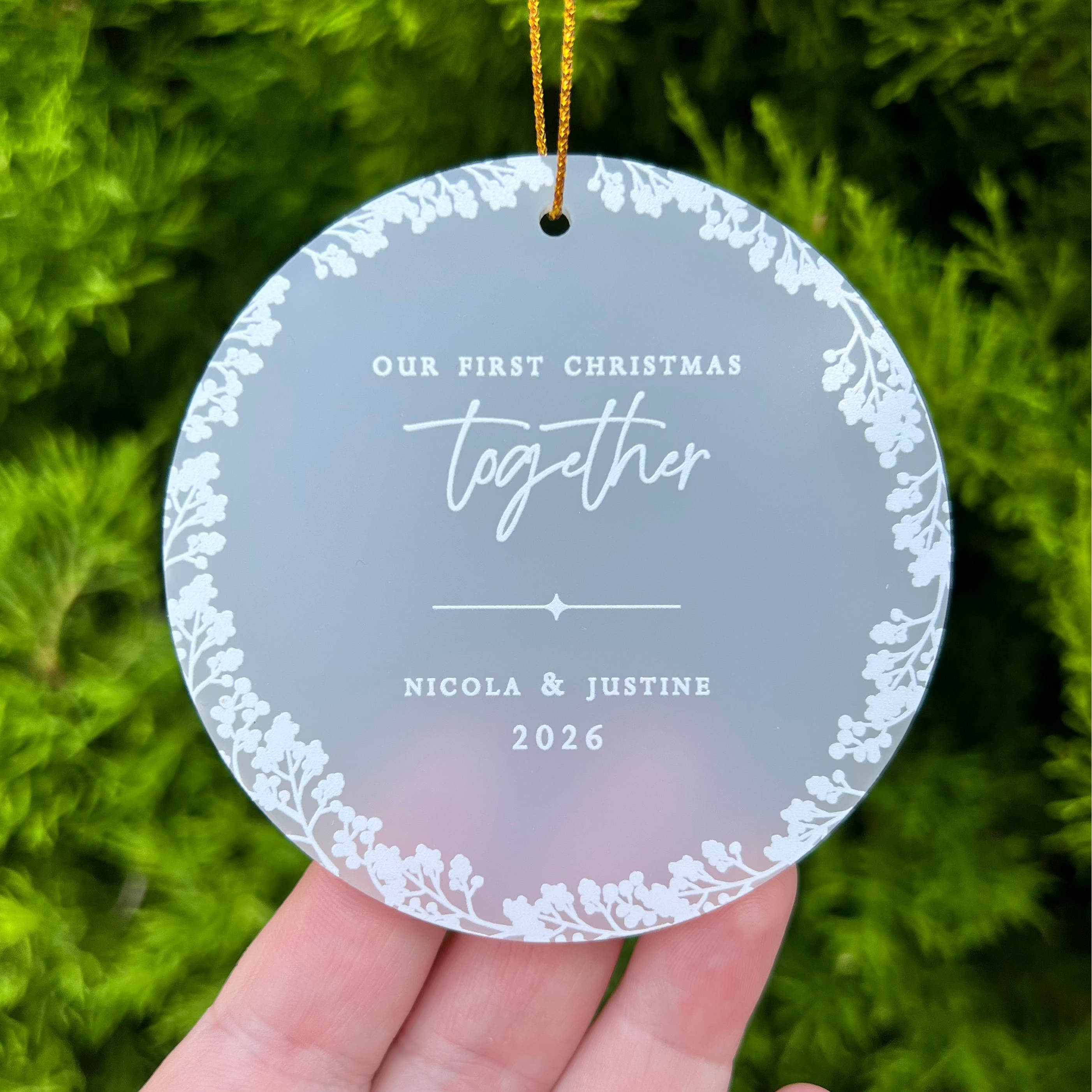 A personalized round acrylic Christmas ornament held in a hand, featuring a frosted gray center with white floral borders and the text 'Our First Christmas Together Nicola & Justine 2026', set against a blurred green background.