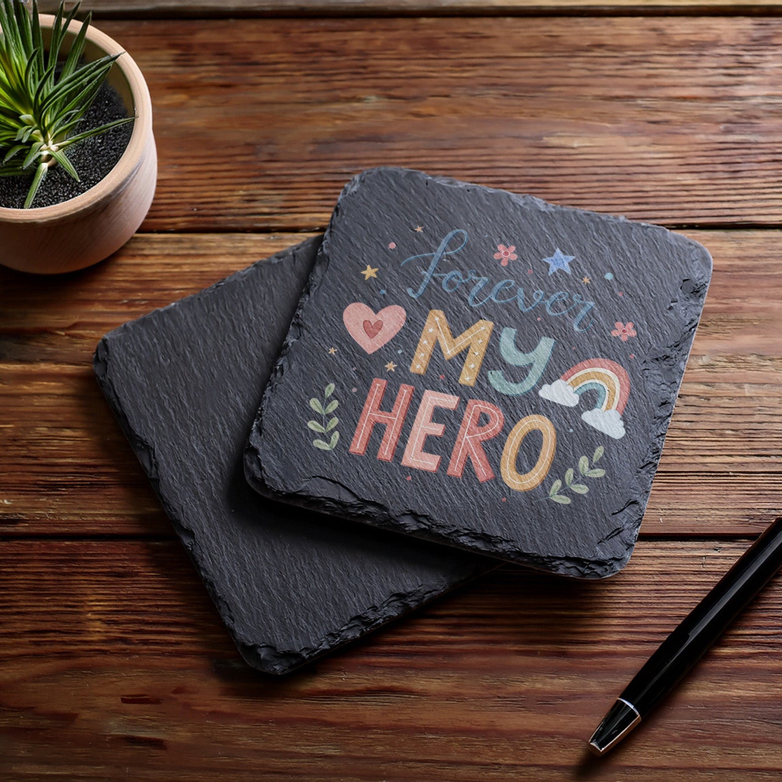 Slate coaster with 'Forever My Hero' text on a wooden surface with a plant and pen.