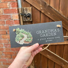 Hand holding a sign that reads 'Grandma's Garden' in front of a wooden door and brick wall.