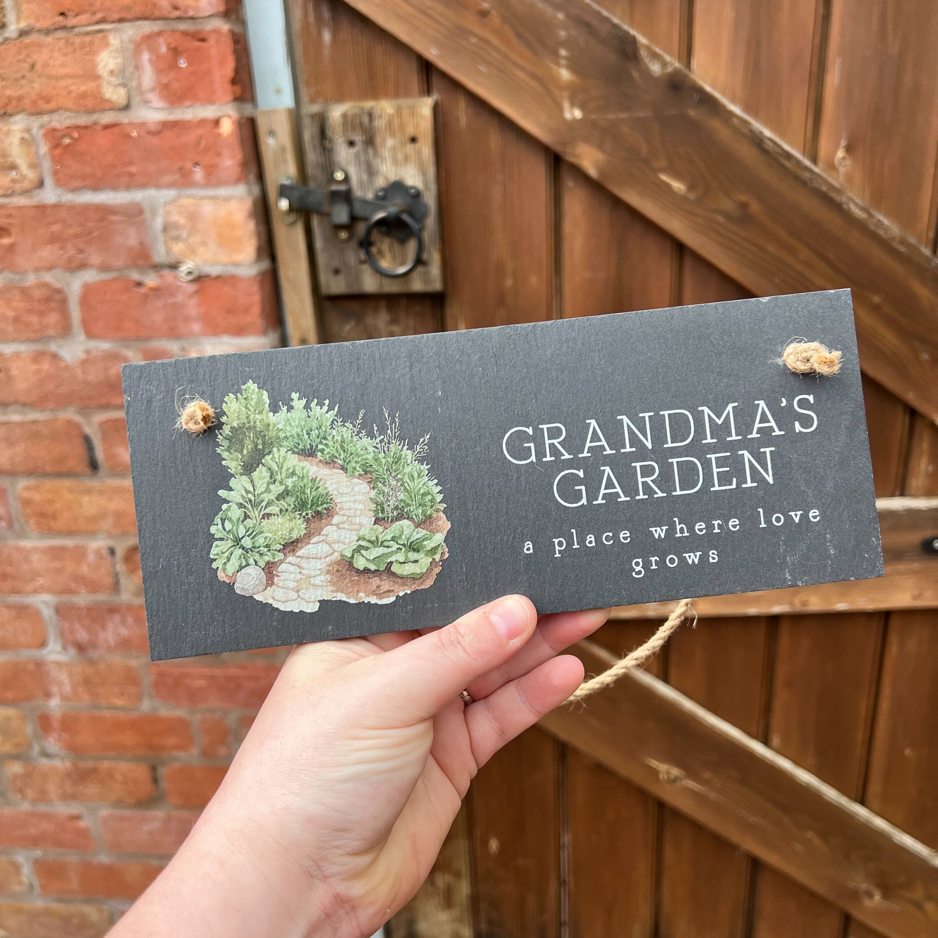 Hand holding a sign that reads 'Grandma's Garden' in front of a wooden door and brick wall.