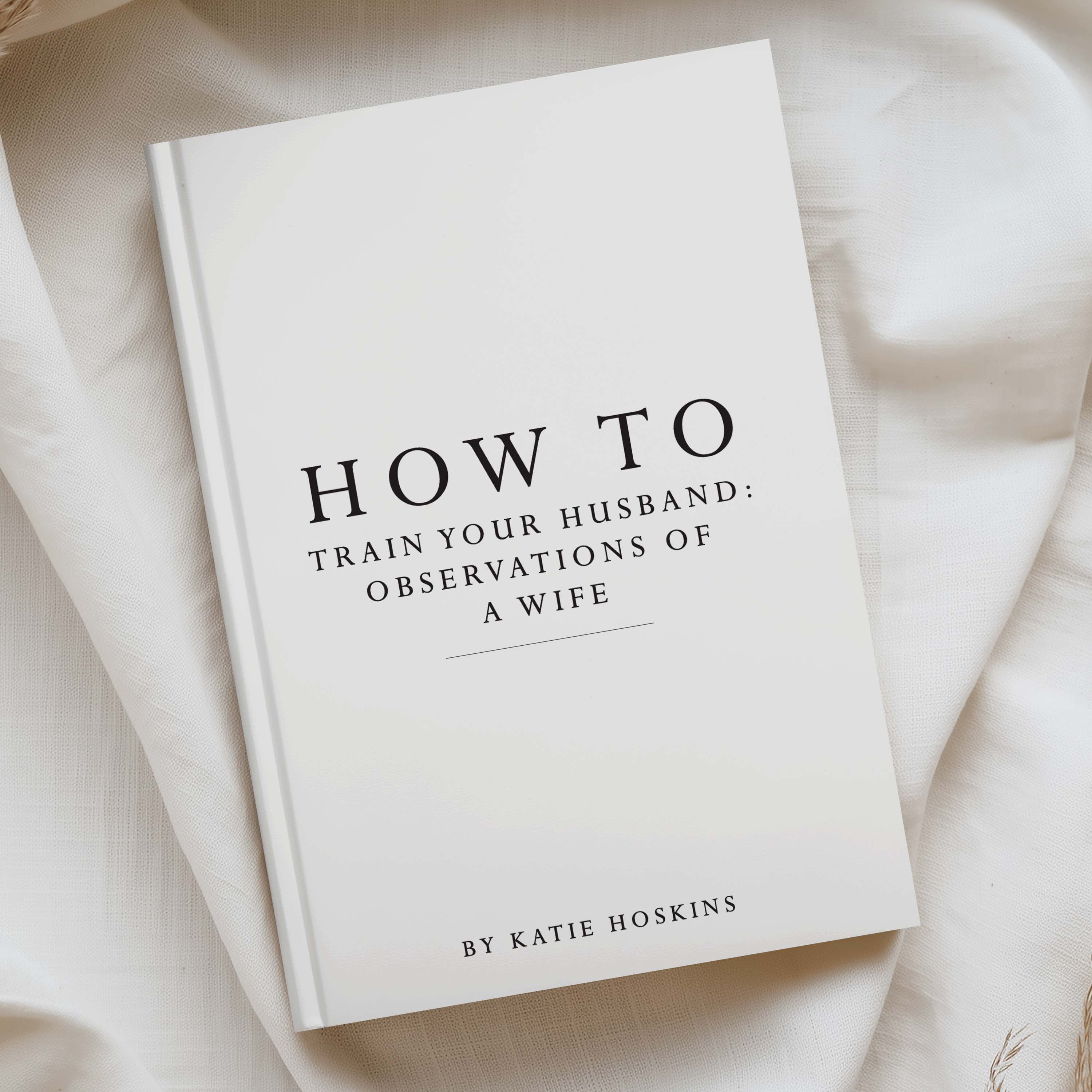 A hardcover book titled 'HOW TO TRAIN YOUR HUSBAND: OBSERVATIONS OF A WIFE' by Katie Hoskins, photographed on draped cream fabric.