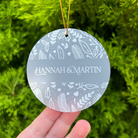 A round frosted glass Christmas ornament with white botanical patterns and the personalized name 'HANNAH & MARTIN' in the center, hanging from a gold string and held by a hand against a green pine background.