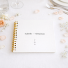white wedding guest book with gold binder on white table cloth with glassware and pink flower petals