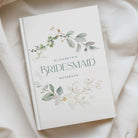 A personalised bridesmaid notebook titled 'Elizabeth's Bridesmaid Notebook' with watercolour eucalyptus and floral illustrations on a white cover, styled on draped cream fabric.