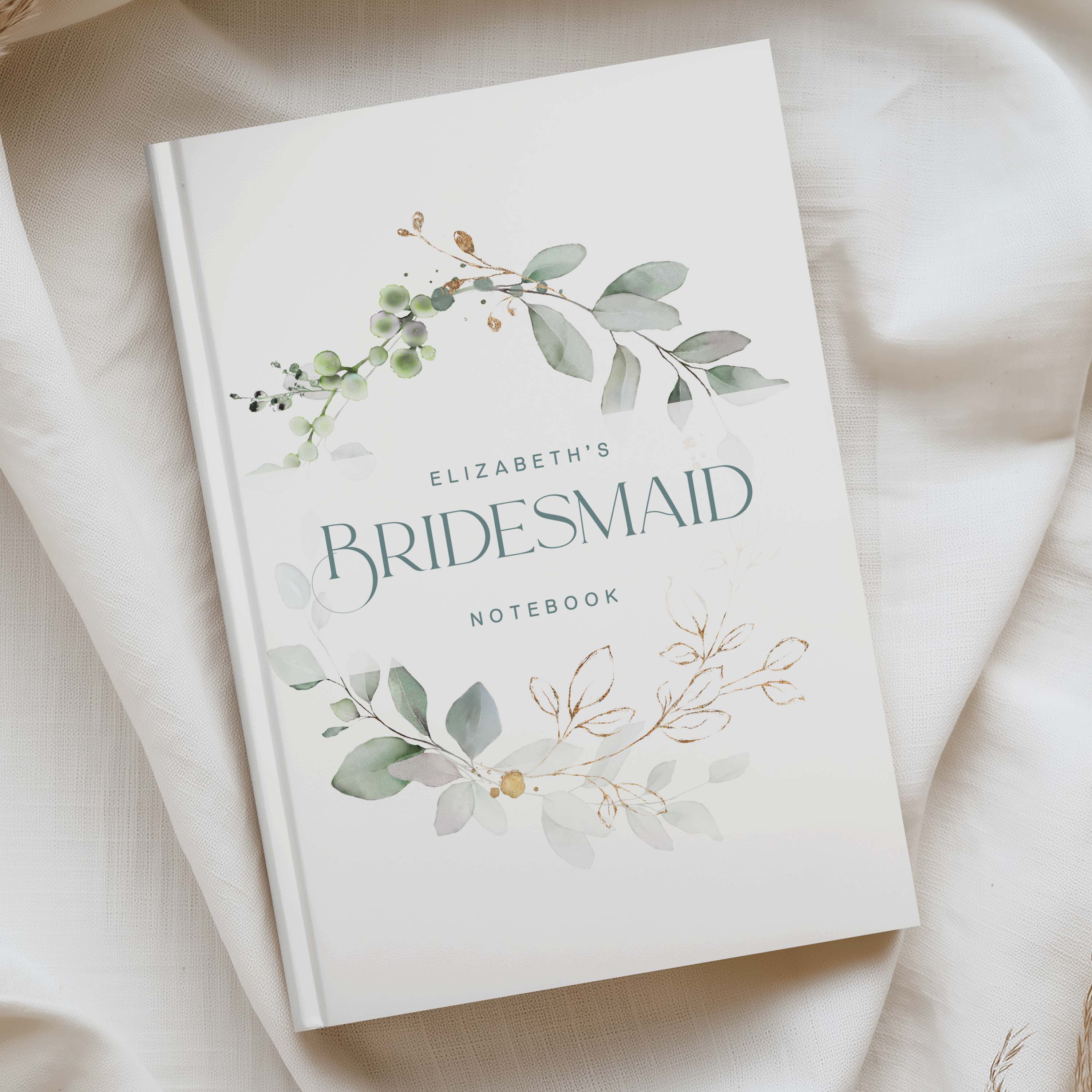 A personalised bridesmaid notebook titled 'Elizabeth's Bridesmaid Notebook' with watercolour eucalyptus and floral illustrations on a white cover, styled on draped cream fabric.