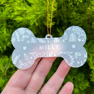 A bone-shaped acrylic pet ornament with the name 'MILLY' in the center, held in a hand against a background of green pine branches. The ornament is frosted with white leaf and paw print patterns and hangs from a gold string.