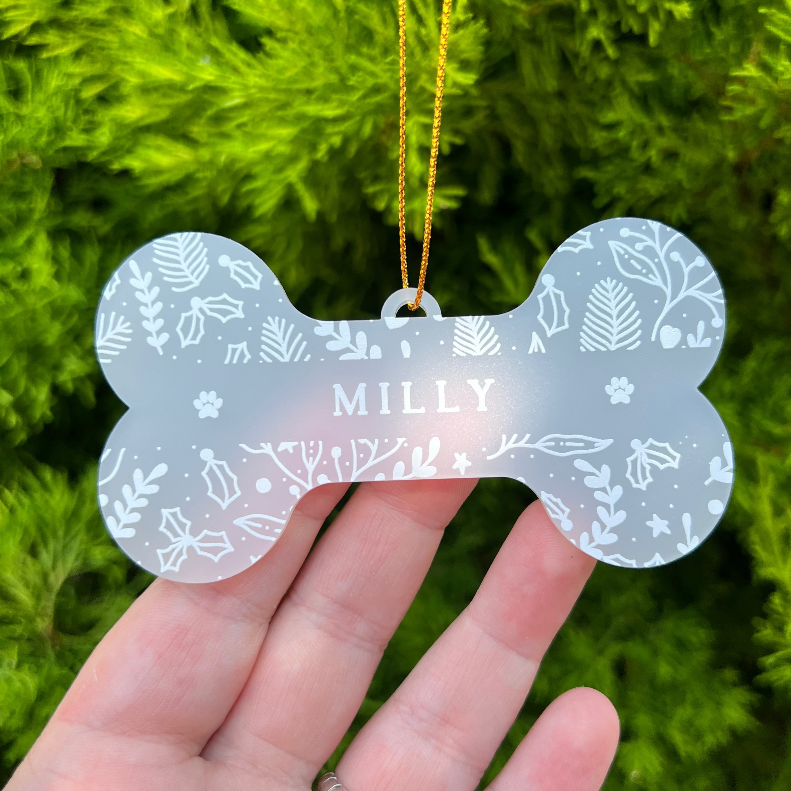A bone-shaped acrylic pet ornament with the name 'MILLY' in the center, held in a hand against a background of green pine branches. The ornament is frosted with white leaf and paw print patterns and hangs from a gold string.