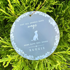 A round frosted acrylic pet memorial hanging ornament with white floral border and text reading 'NOT ALL ANGELS HAVE wings SOME HAVE WET NOSES REGGIE' and a dog silhouette, hanging from a tree branch.