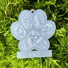 A frosted acrylic paw-shaped ornament with white leaf and heart patterns and the name 'LILY' at the bottom, photographed against bright green foliage.