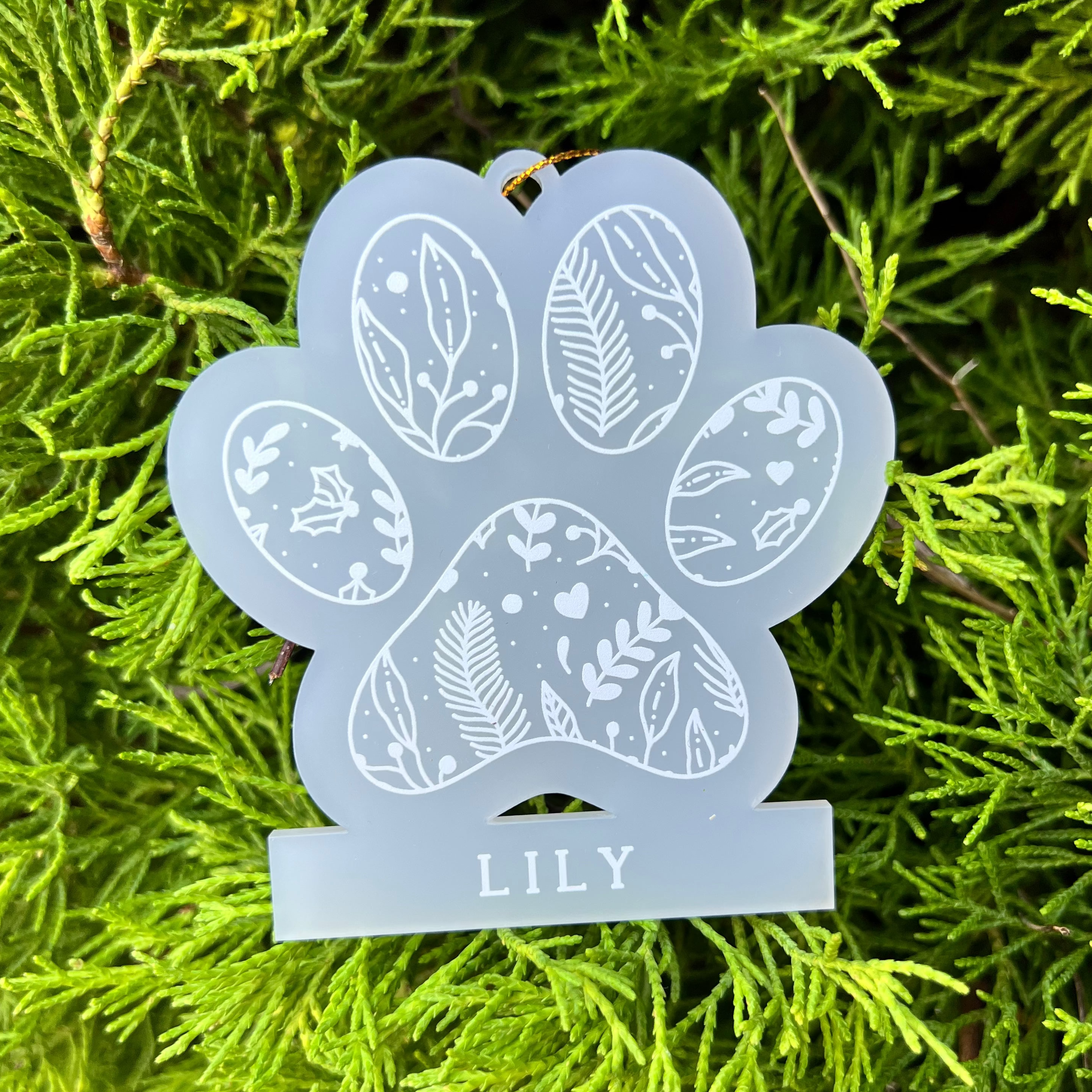 A frosted acrylic paw-shaped ornament with white leaf and heart patterns and the name 'LILY' at the bottom, photographed against bright green foliage.