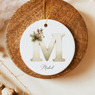 A round white personalized Christmas ornament featuring a large letter 'M' with a small deer and pine sprig illustration, hanging from a gold string on a rustic wooden board.