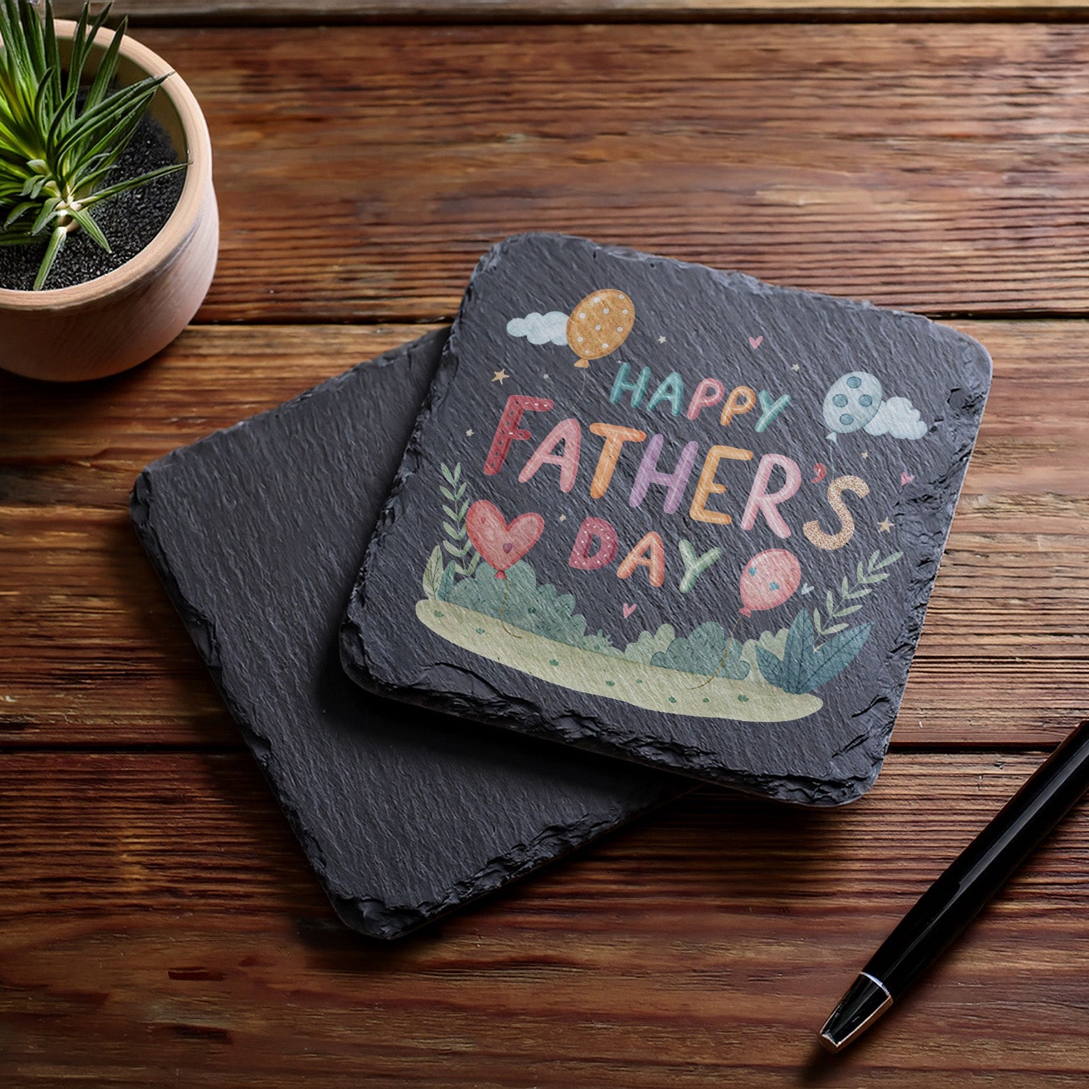 Slate coaster with 'Happy Father's Day' design on a wooden surface with a pen and plant.